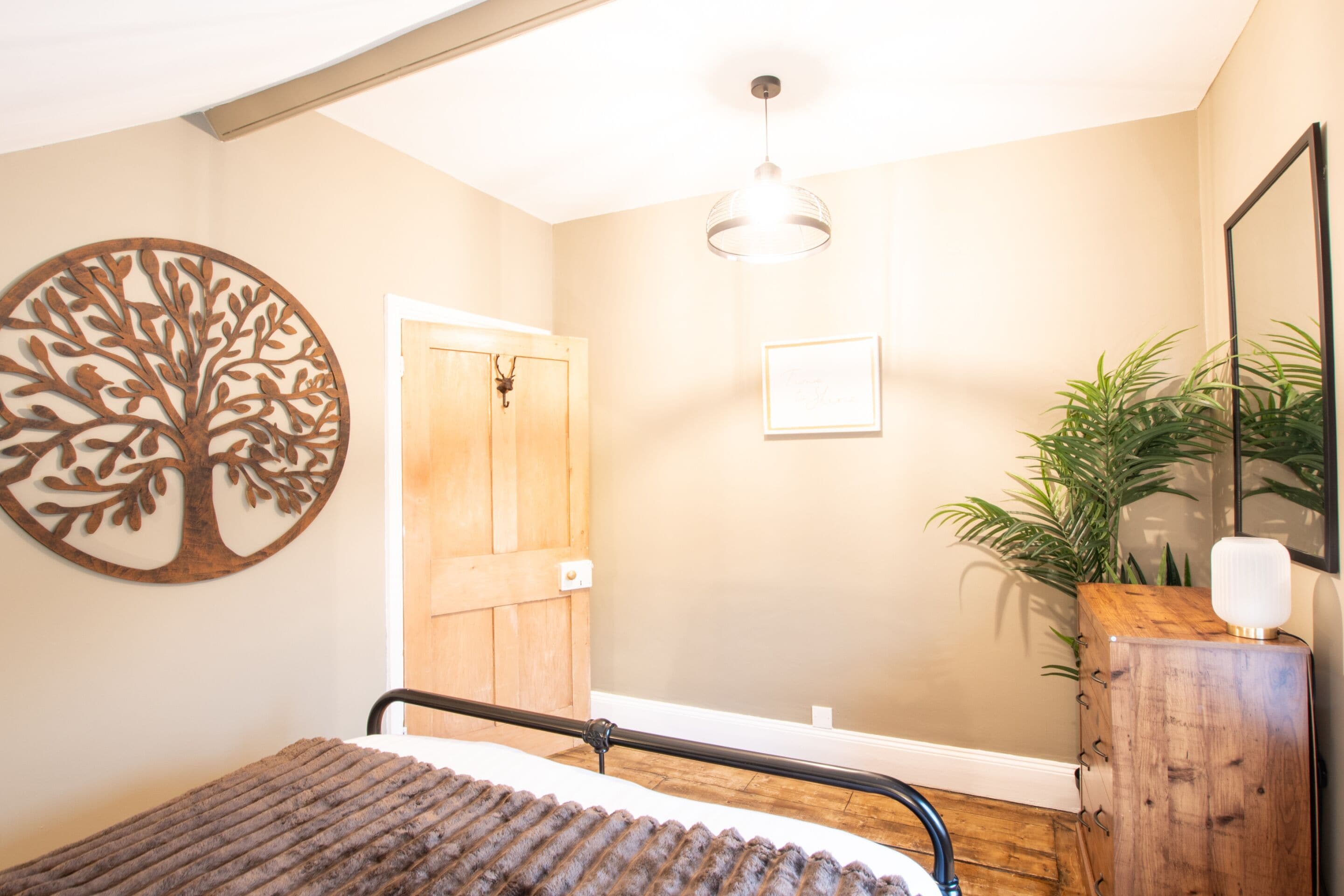A bedroom with a bed, a wooden door, a wall-mounted tree decoration, a hanging light fixture, and a potted plant.
