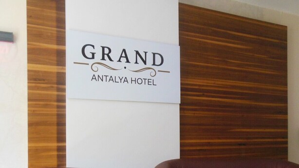 Uma área de recepção com uma placa para o Grand Antalya Hotel, dois sofás de couro marrom e um painel de parede de madeira.