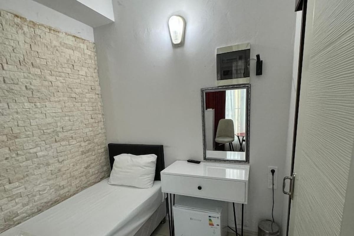 A small, compact bedroom with a single bed, a white dresser, a mirror, and a small white appliance.