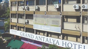 Um edifício de vários andares com inúmeras unidades de ar condicionado, uma placa com o nome Grand Antalya Hotel e um banner promovendo um negócio local.