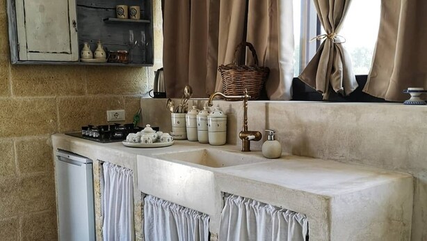 A kitchen with a stone wall, a built-in shelf with kitchenware, a sink, and a window with curtains. - Relais Grottone (Ostuni)