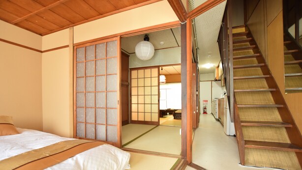 Ein traditioneller japanischer Raum mit Tatami-Böden, einer Schiebetür, einem Bett mit weißer und brauner Bettdecke sowie einer Holztreppe.