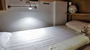 Un lit simple avec une tête de lit, une table de chevet, une lampe fixée au mur, une prise de courant et un ventilateur de plafond.