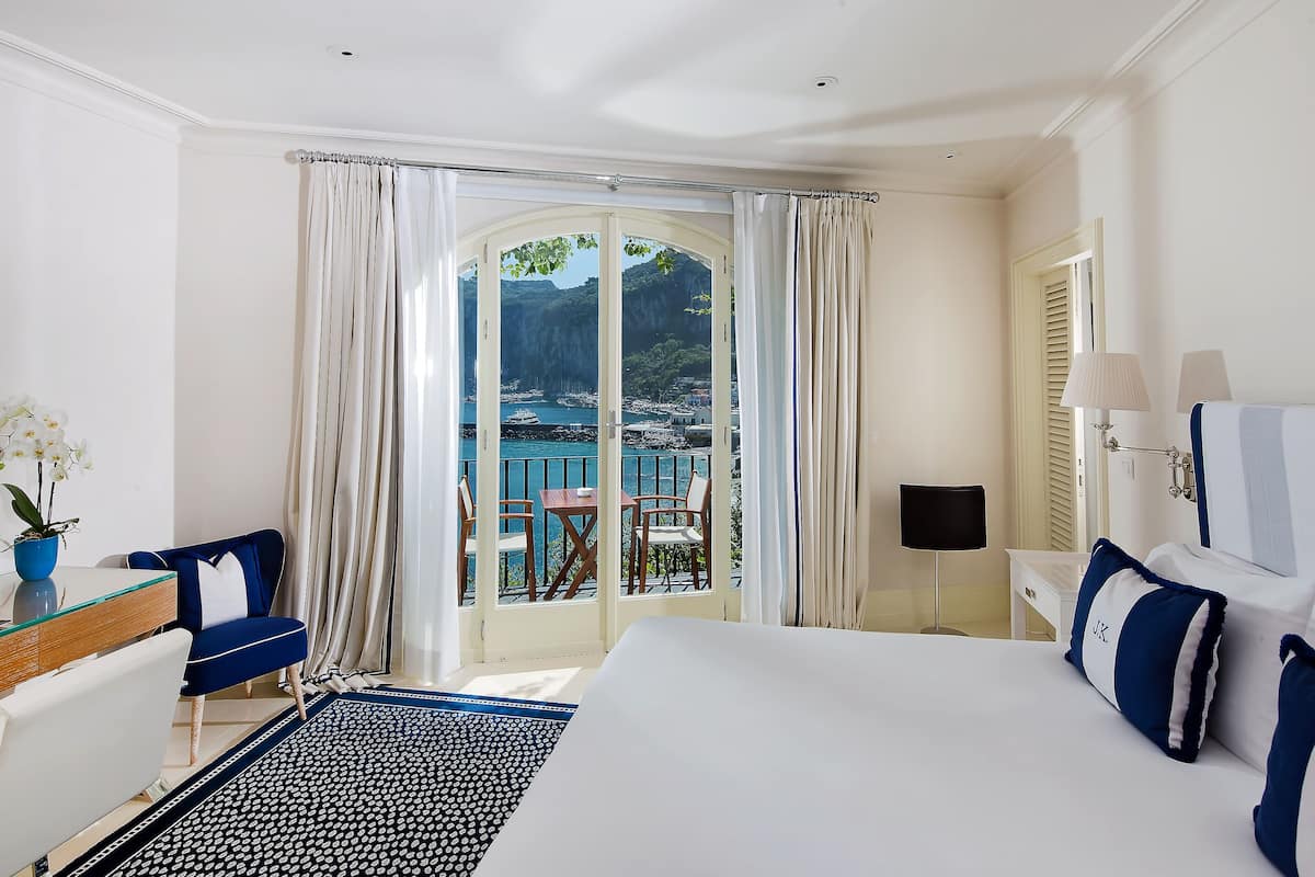 A hotel room with a large bed, a desk with a chair, a small table with chairs, and a view of the sea through a glass door.