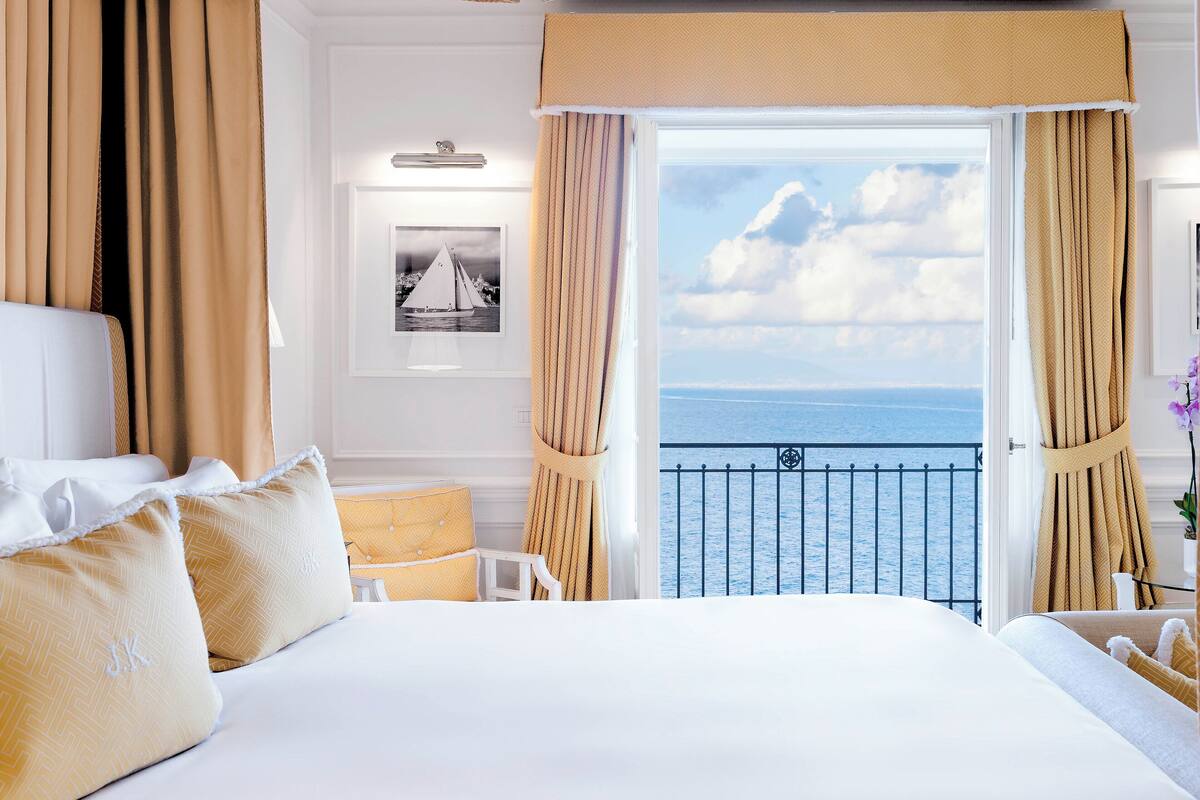 A hotel room with a large bed, a view of the sea, and a balcony with curtains.