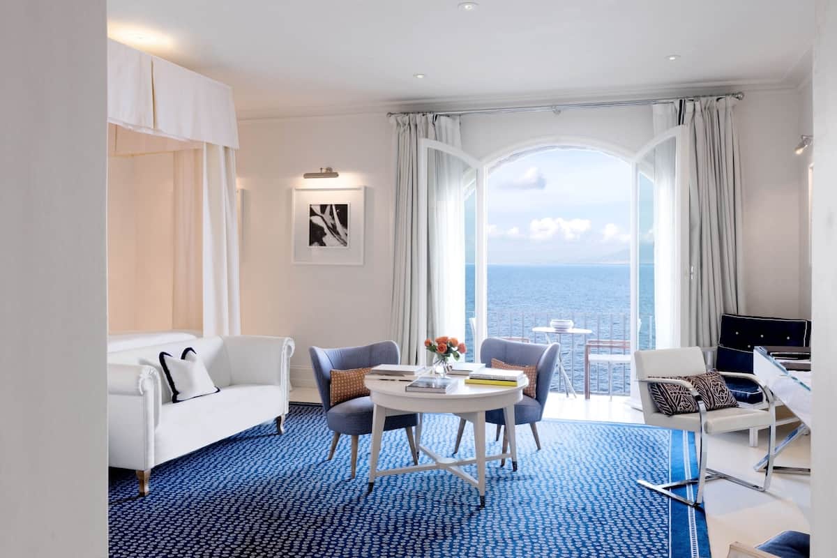 A modern hotel room with a white sofa, two armchairs, a small round table with books and flowers, a desk with a chair, and a large window offering a view of the sea.