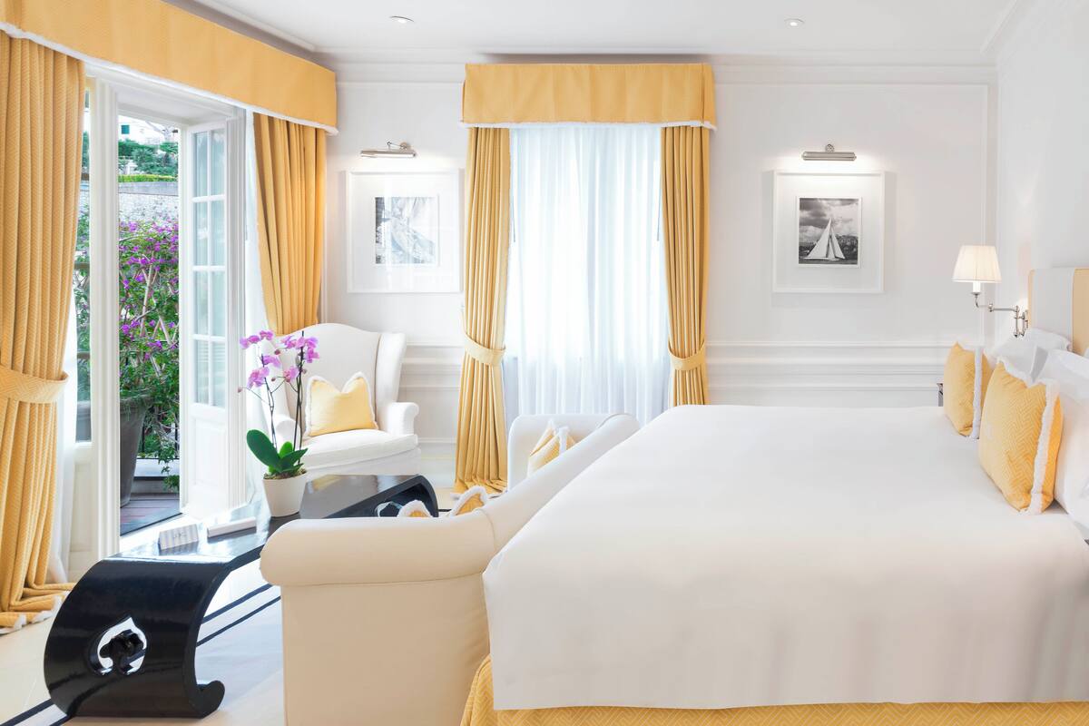 A hotel room with a large bed, two white armchairs, a small table with a potted plant, and a view of the outdoors through a glass door.