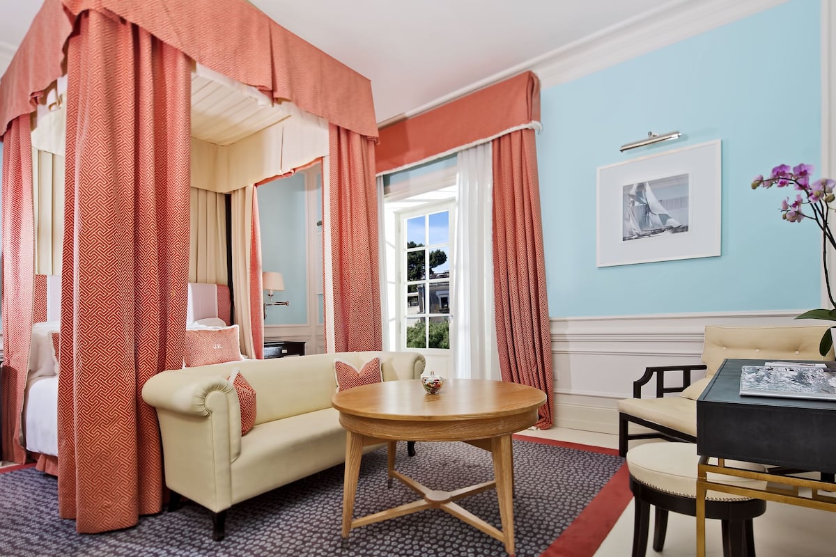 A room with a bed, a round wooden table, a sofa, and a chair. There is a window with curtains, a framed picture on the wall, and a vase with flowers.