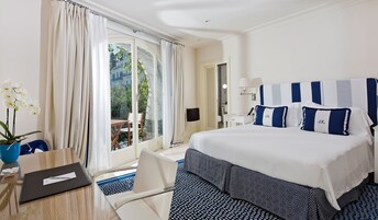 A hotel room with a large bed, a desk with a chair, a potted plant, and a view of the outdoors.