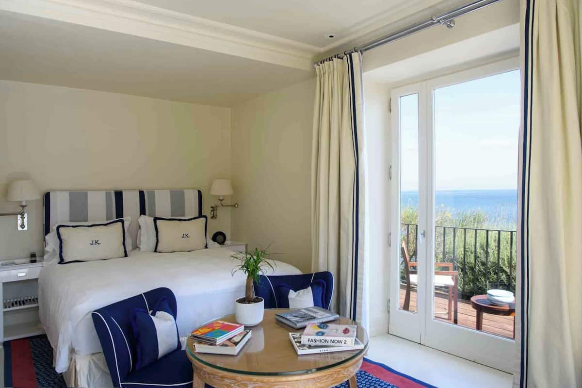 A hotel room with a bed, bedside tables, a round glass table with books, a potted plant, and a balcony with a view of the sea.