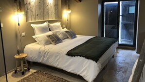 Ein Schlafzimmer mit einem großen Bett, zwei Wandleuchten, einem hölzernen Hocker und Blick ins Freie durch eine Tür.
