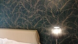 Un lit avec du linge de lit blanc, une table de chevet ronde avec une lampe, et un mur orné d’un motif de feuilles.