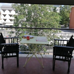 Un balcón con dos sillas negras, una mesita con una taza roja, y vista a un edificio con un letrero que dice 'GENERATIF'.