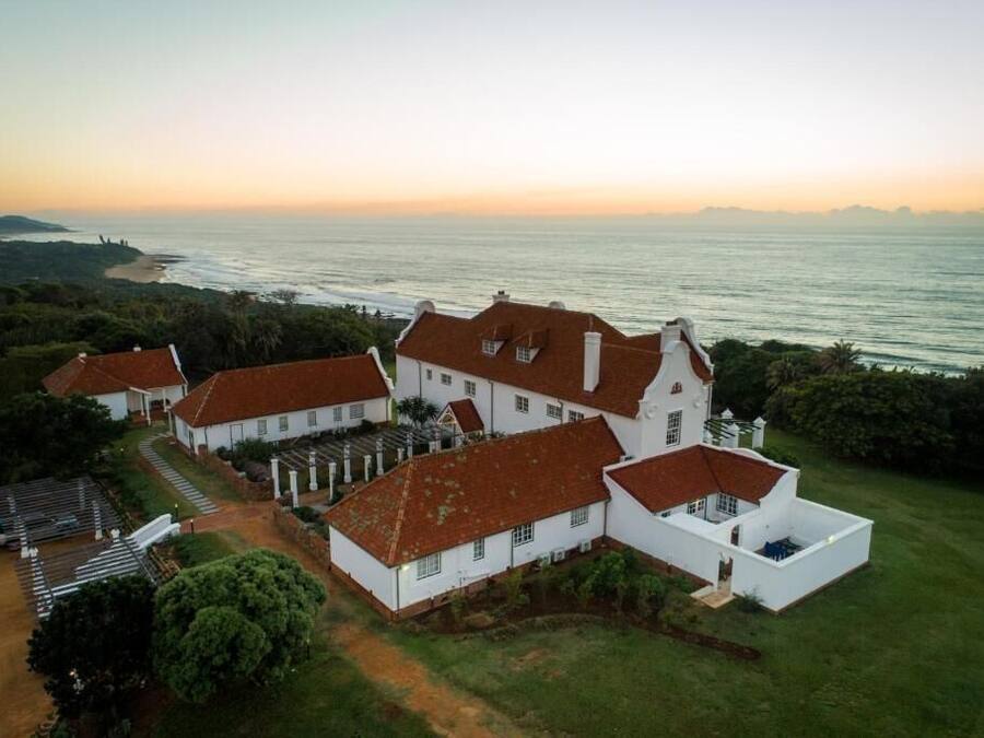 Un edificio grande de color blanco con techos rojos, rodeado de vegetación y con vistas a un cuerpo de agua.