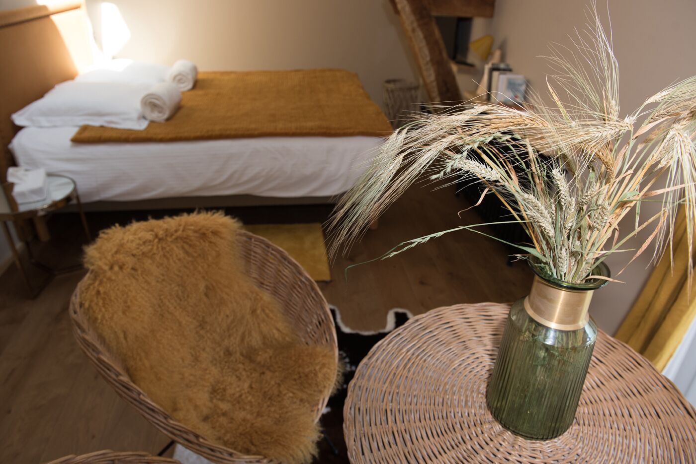 A room with a bed, a wicker chair, and a vase with dried plants.