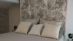 Un dormitorio con cama, almohadas, cabecera con estampado de hojas tropicales, una lámpara de pared y una mesita con silla.