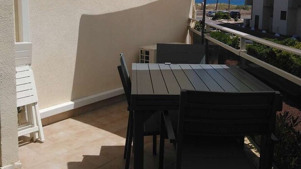Un patio exterior con mesa y sillas, un toldo blanco y vista al mar y edificios al fondo.