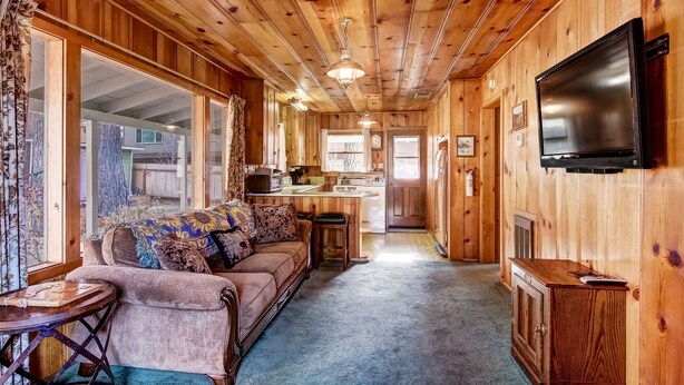 A cozy cabin interior with a wooden interior, a sofa, a small round table, and a kitchen area visible through an open door. - 3993c Cedar Lake Tahoe 2 Bedroom Cabin by RedAwning (South Lake Tahoe)