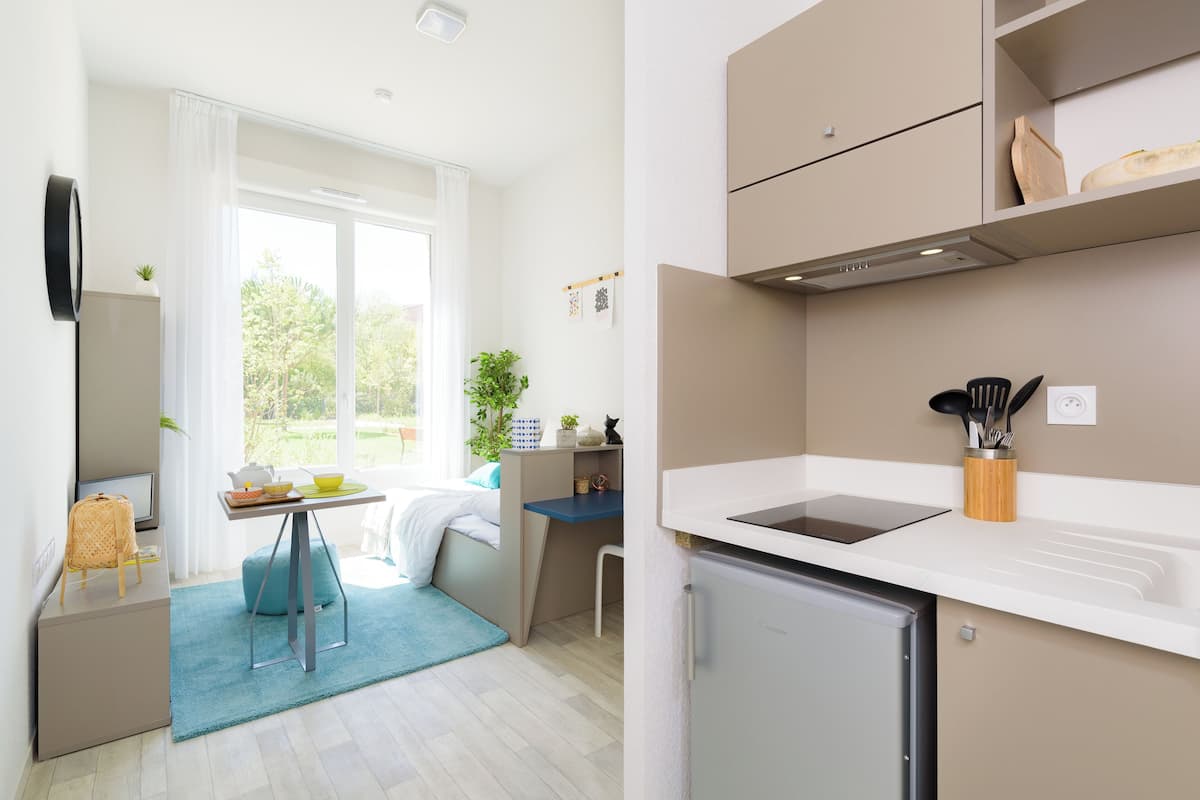 Eine kompakte Küche mit integriertem Kühlschrank, ein kleiner Essbereich mit blauem Teppich und ein Fenster, das für natürliches Licht sorgt.