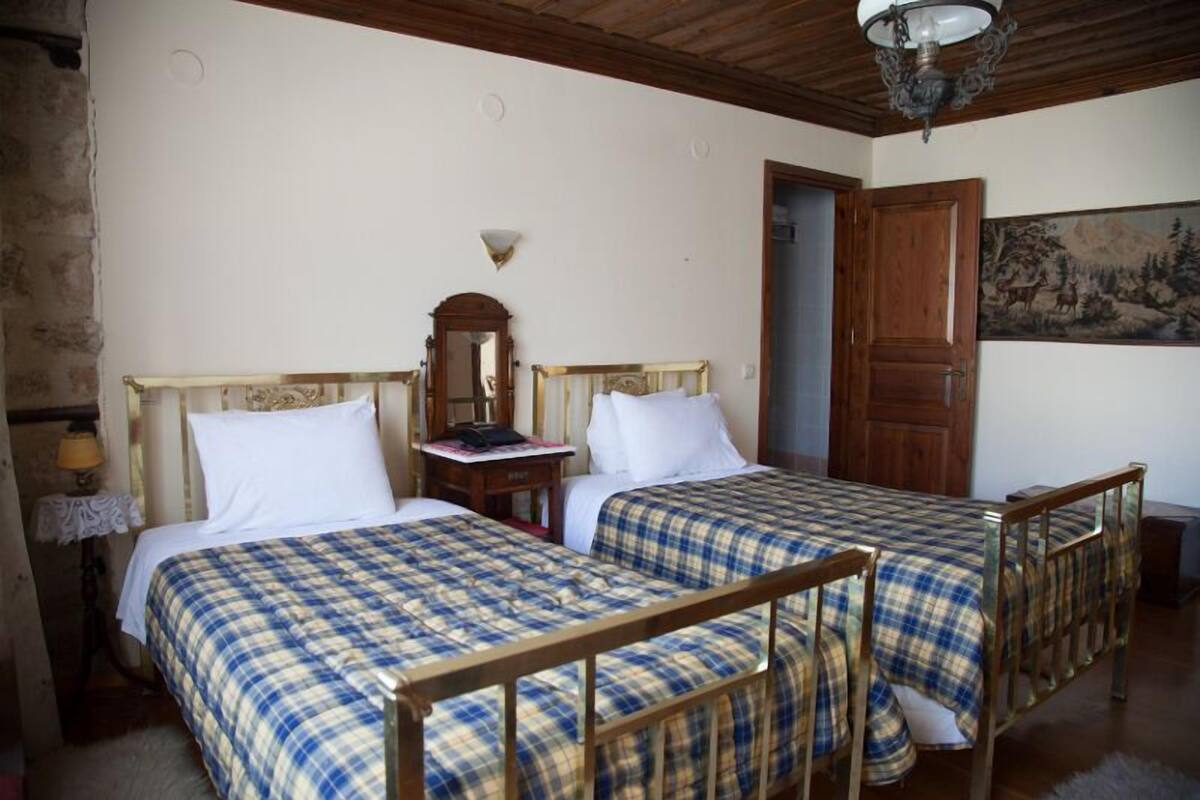 A room with two beds, each with a wooden frame and plaid bedding. There is a wooden door, a wall-mounted lamp, and a painting on the wall.