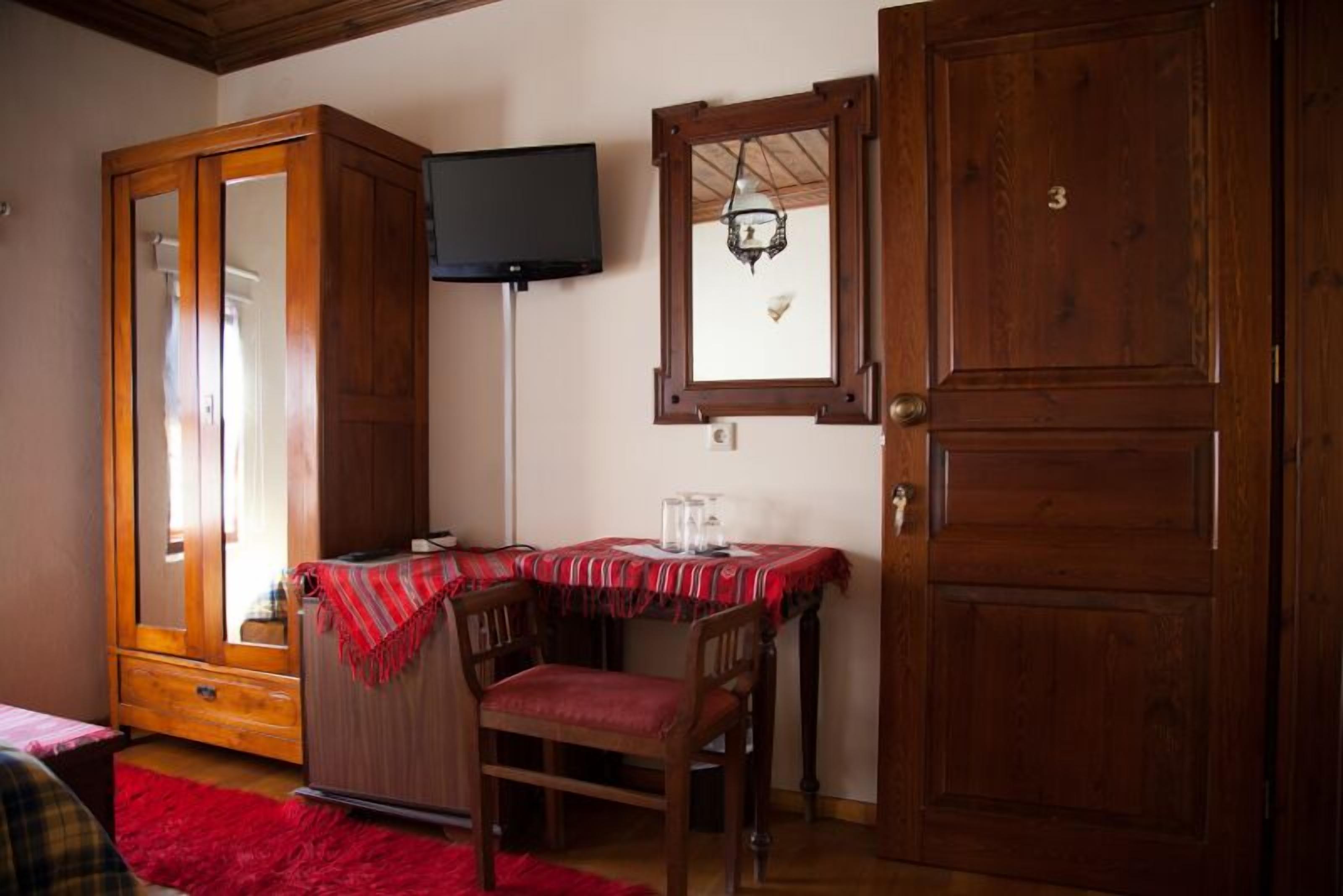 Una habitación con un armario de madera, una mesa con mantel rojo, una silla, un televisor montado en la pared y una ventana con lámpara colgante.