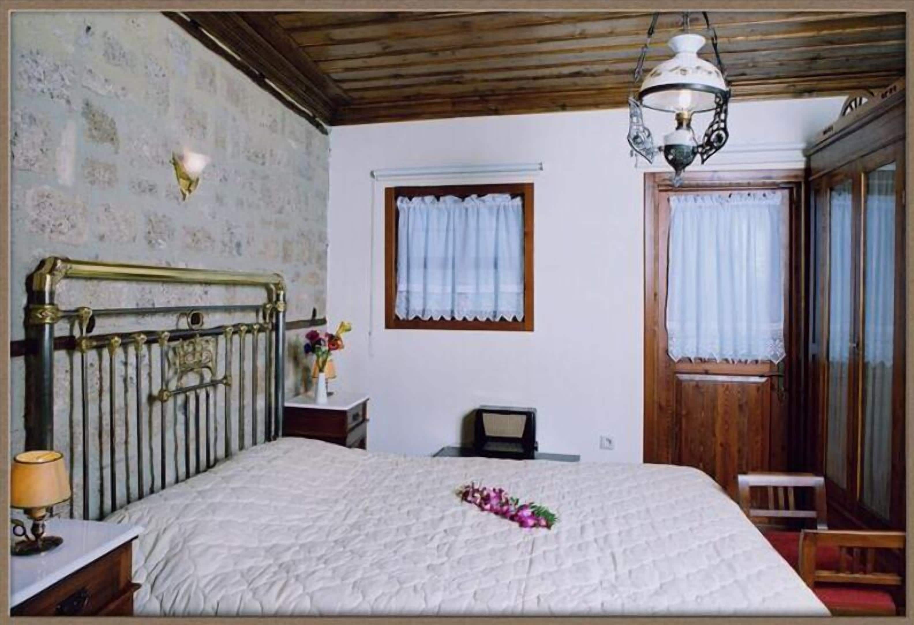 A bedroom with a bed, a wooden headboard, a nightstand, a lamp, a window with curtains, and a wooden door.