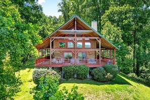 A large log cabin with a porch and multiple windows, surrounded by lush greenery.