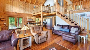 Un intérieur spacieux en bois, avec un plafond haut, un escalier, un coin cuisine et un espace salon agrémenté de canapés en cuir et d’une table basse.