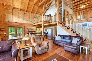 A spacious wooden interior with a high ceiling, a staircase, a kitchen area, and a seating area with leather sofas and a coffee table.