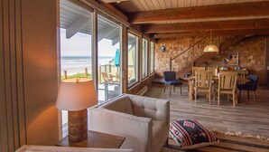 A cozy interior with a wooden ceiling, large windows overlooking the beach, a dining area with wooden furniture, and a staircase leading to an upper level. - Ocean Front on Miles of Sandy Beach! Dog Friendly! (Yachats)
