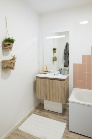 A modern bathroom with a wooden vanity, white sink, and a large mirror. There are two wicker baskets on the wall, a white rug on the floor, and a bathtub with a beige tile wall. - Residence Sweetly St Augustin (Bordeaux)