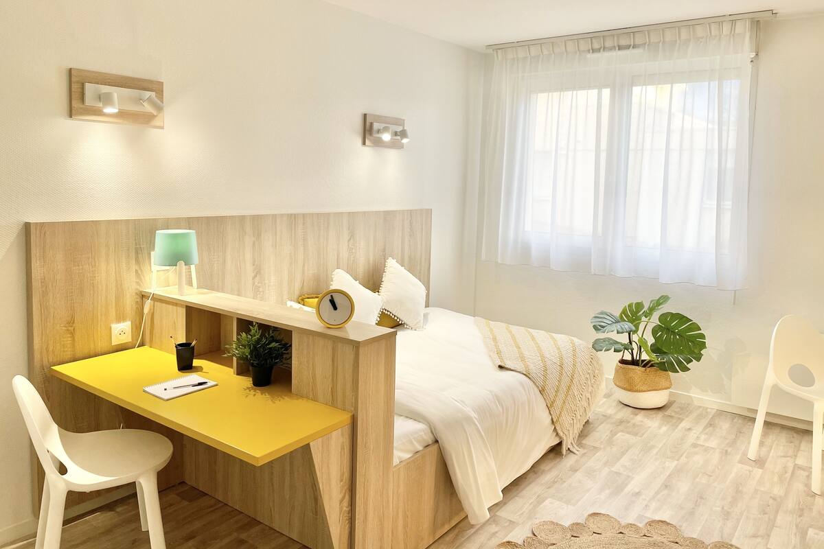 Een moderne slaapkamer met een houten hoofdbord, een geel bureau met een lamp, een bed met witte beddengoed, een raam met dunne gordijnen en een potplant.