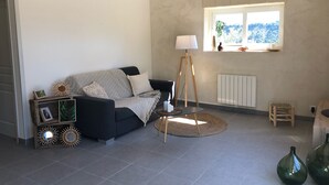 A living room with a dark sofa, a floor lamp, a small table, and a window with a view of greenery. - Mas provençal La Serallère in the heart of olive groves (Venterol)