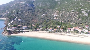 Vue aérienne d’une zone côtière avec une plage, des maisons et un paysage montagneux en arrière-plan.