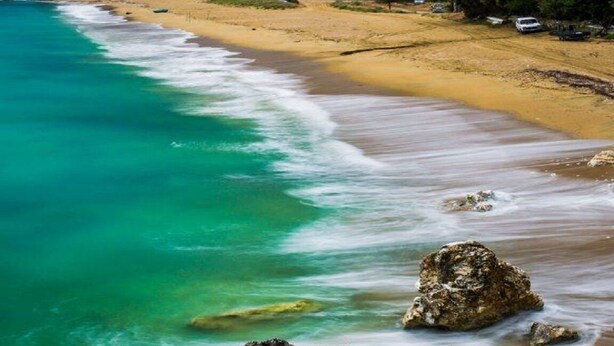 Ein Strand mit klarem türkisblauem Wasser, einem Sandufer und Häusern, die sich einen Hügel hinaufziehen.