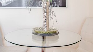 Una mesa de vidrio redonda con un florero de flores blancas y hojas verdes, flanqueada por dos sillas blancas. Se distingue un paisaje urbano enmarcado al fondo.
