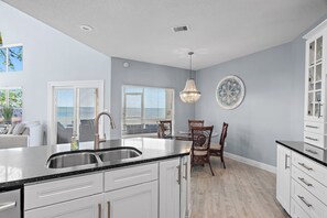 Una cocina con armarios blancos, encimera negra, pileta de acero inoxidable, comedor con sillas de madera y un plato decorativo en la pared.