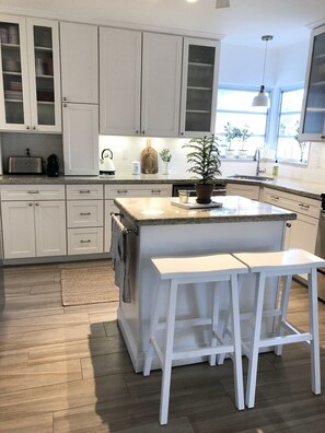 Une cuisine moderne avec des meubles blancs, un comptoir en granit, un îlot central doté de tabourets de bar et une vue sur l’extérieur grâce à de grandes fenêtres.