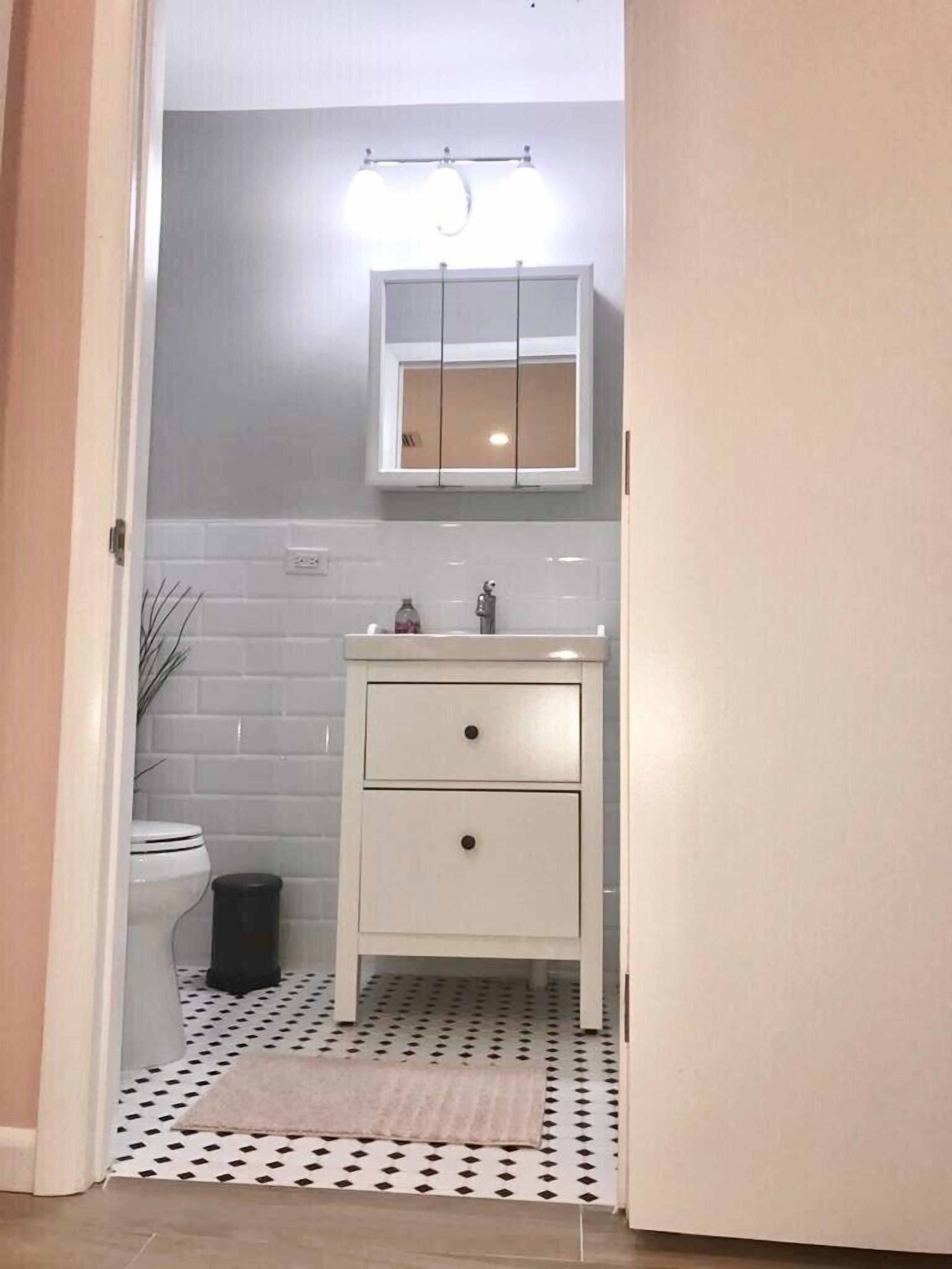 Un baño con un lavamanos blanco, dos cajones, un espejo sobre el lavamanos y un cubo de basura negro.