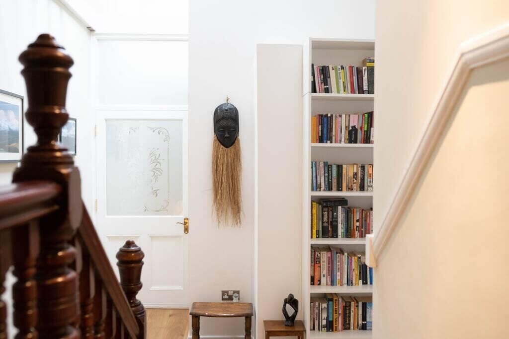 Eine Treppe mit Holzgeländer führt zu einem Raum, der eine weiße Tür, ein Bücherregal voller Bücher und eine dekorative Maske an der Wand hat.