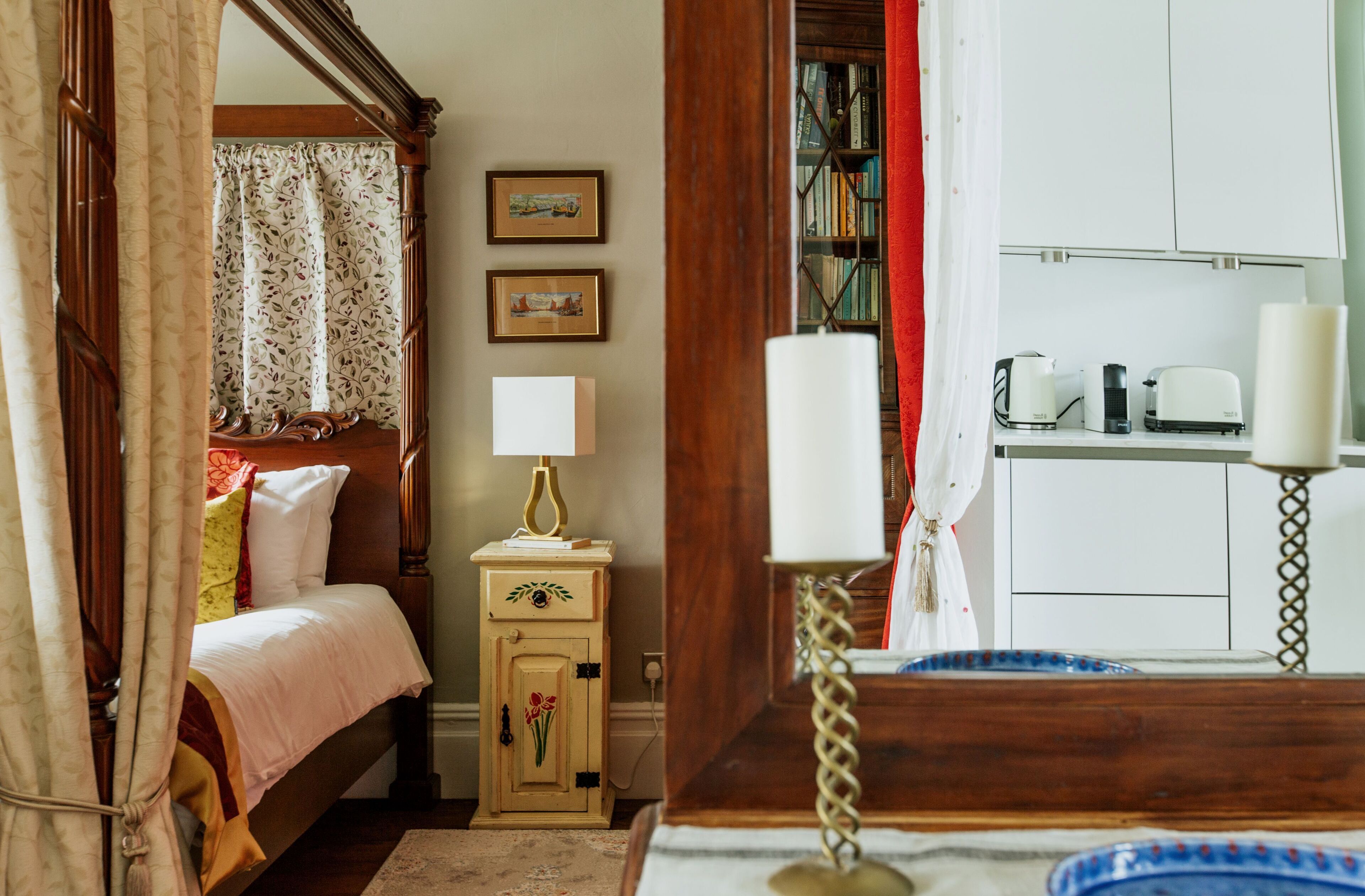A bedroom with a wooden four-poster bed, a bedside table with a lamp, and a small cabinet. There is a bookshelf with books and a red curtain partially covering the window.