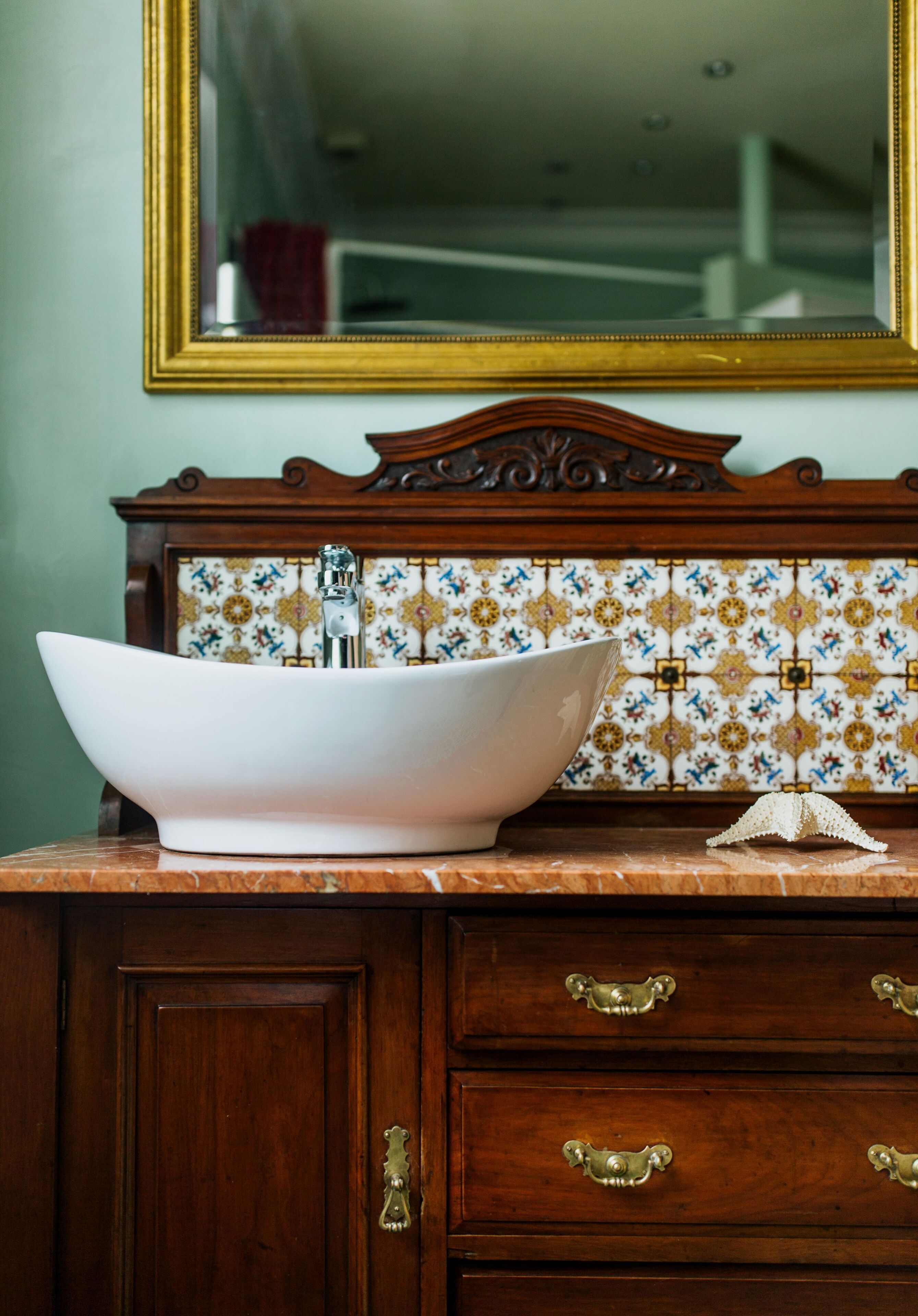 Une salle de bain dotée d’un grand lavabo sur pied blanc, d’un miroir avec un cadre doré et d’un pare-feu en carreaux.