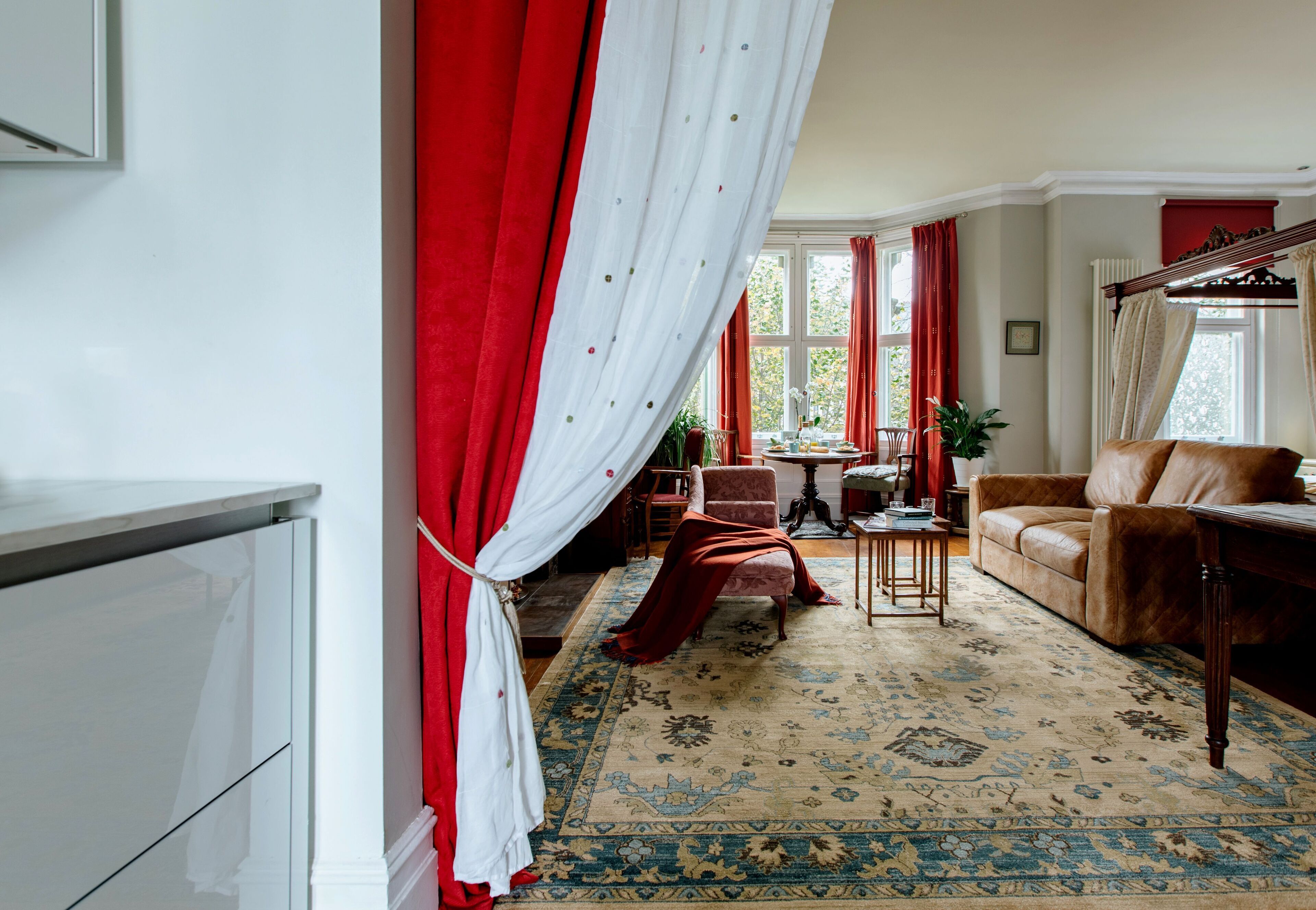 A living room with a large window, a brown leather sofa, a patterned rug, and a red curtain.