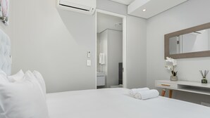 A modern hotel room with a large bed, a bedside table with a mirror, a small desk, and a bathroom visible through an open door.