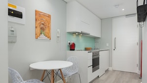 A compact kitchen with white cabinets, a stove, and a sink. There is a white table with two chairs and a painting on the wall.
