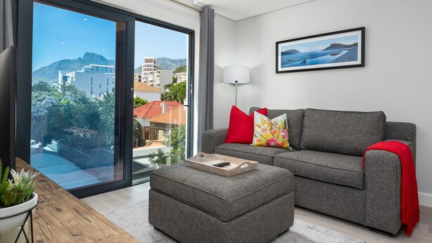 A modern living room with a grey sofa, red and colorful cushions, a wooden coffee table with a tray, a floor lamp, and a large window offering a city view.