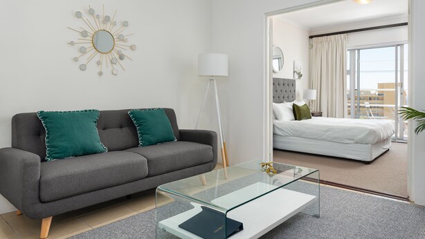 Une chambre d’hôtel moderne avec un canapé gris, une table basse en verre, un grand miroir et un lit avec une literie blanche.