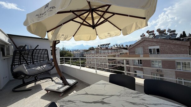 Una terraza en la azotea con mesa, sillas, un parasol, una silla de mimbre y vistas a edificios y montañas.