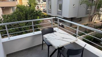 A balcony with a marble table and chairs, a metal railing, and a view of neighboring buildings and greenery.