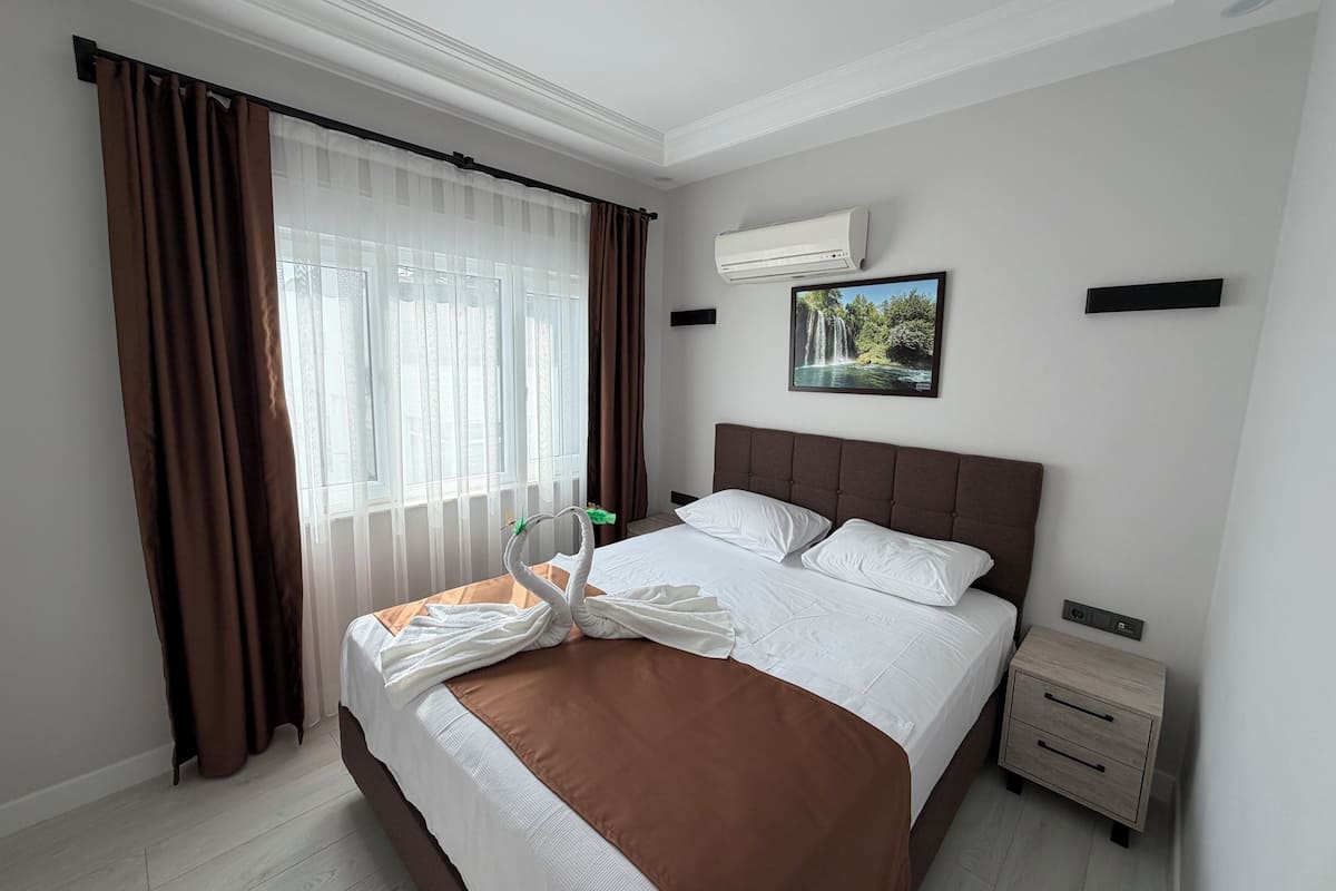 Ein Hotelzimmer mit einem großen Bett, braunen Vorhängen, einer Klimaanlage, einem gerahmten Bild an der Wand und einem Nachttisch.